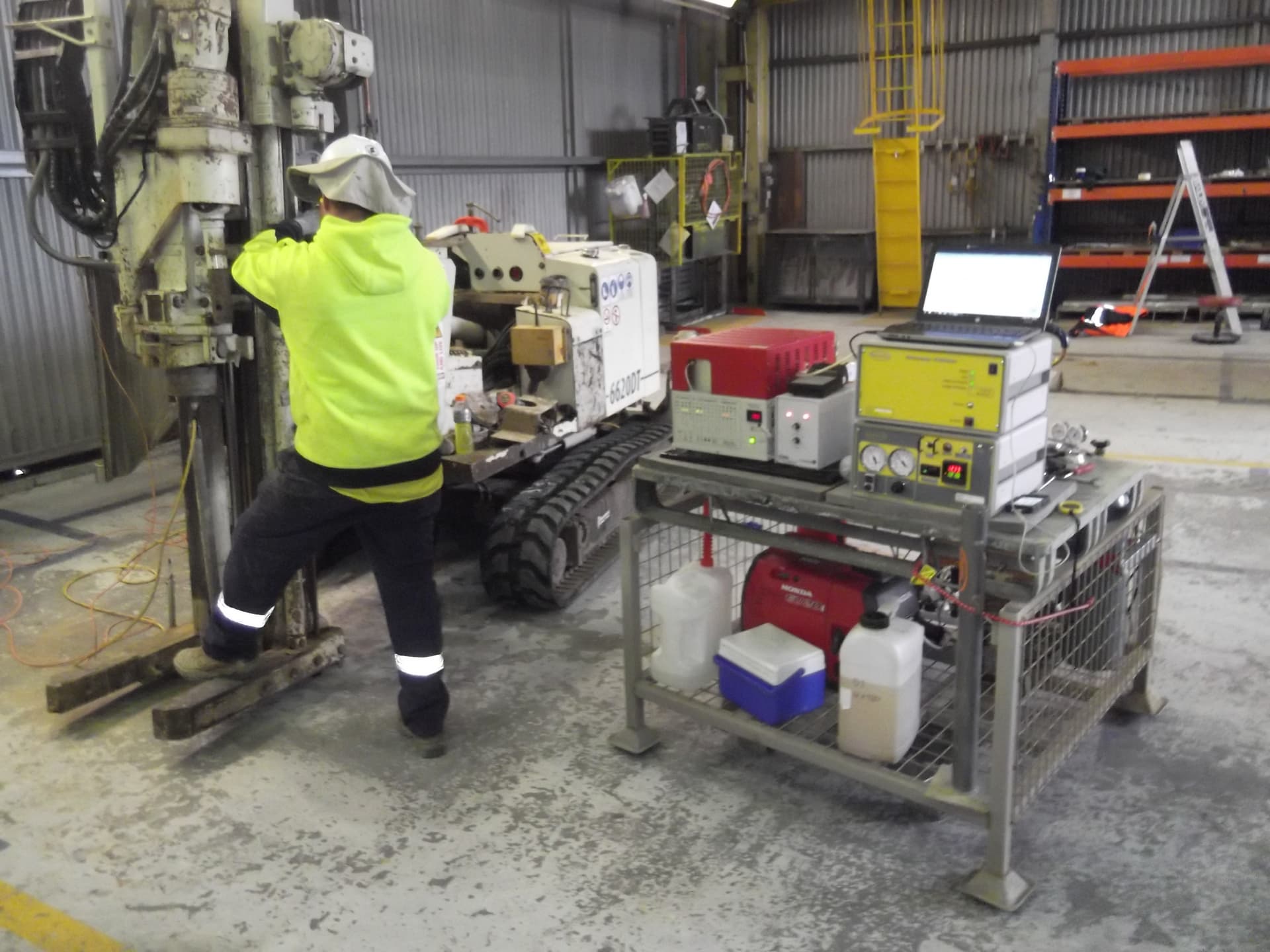 A tracked direct-push rig working indoors under a low industrial roof, with the acquisition equipment racked alongside