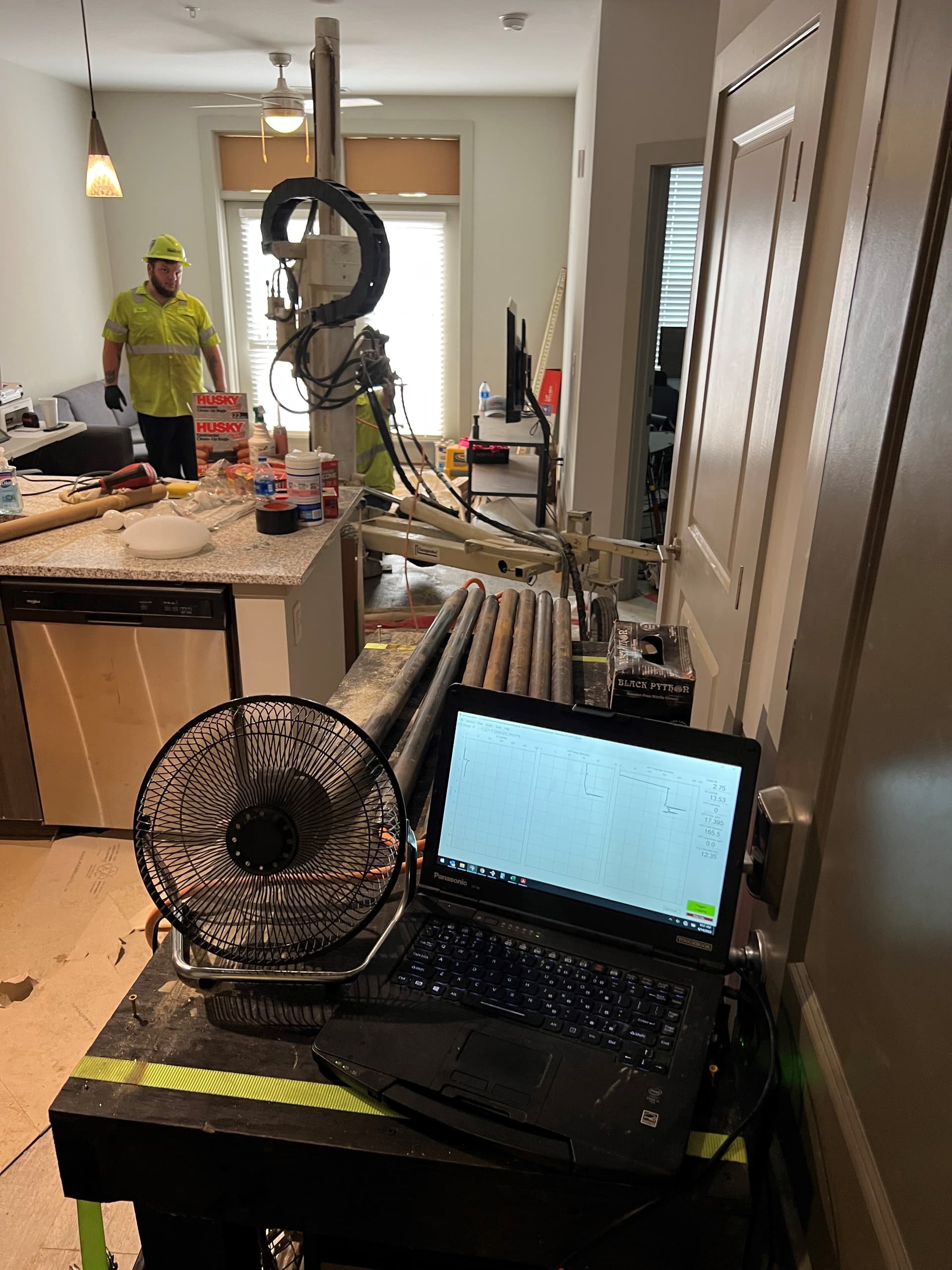 A compact direct-push rig set up inside a finished residential interior, its mast reaching the ceiling, with the logging laptop running on a table beside it